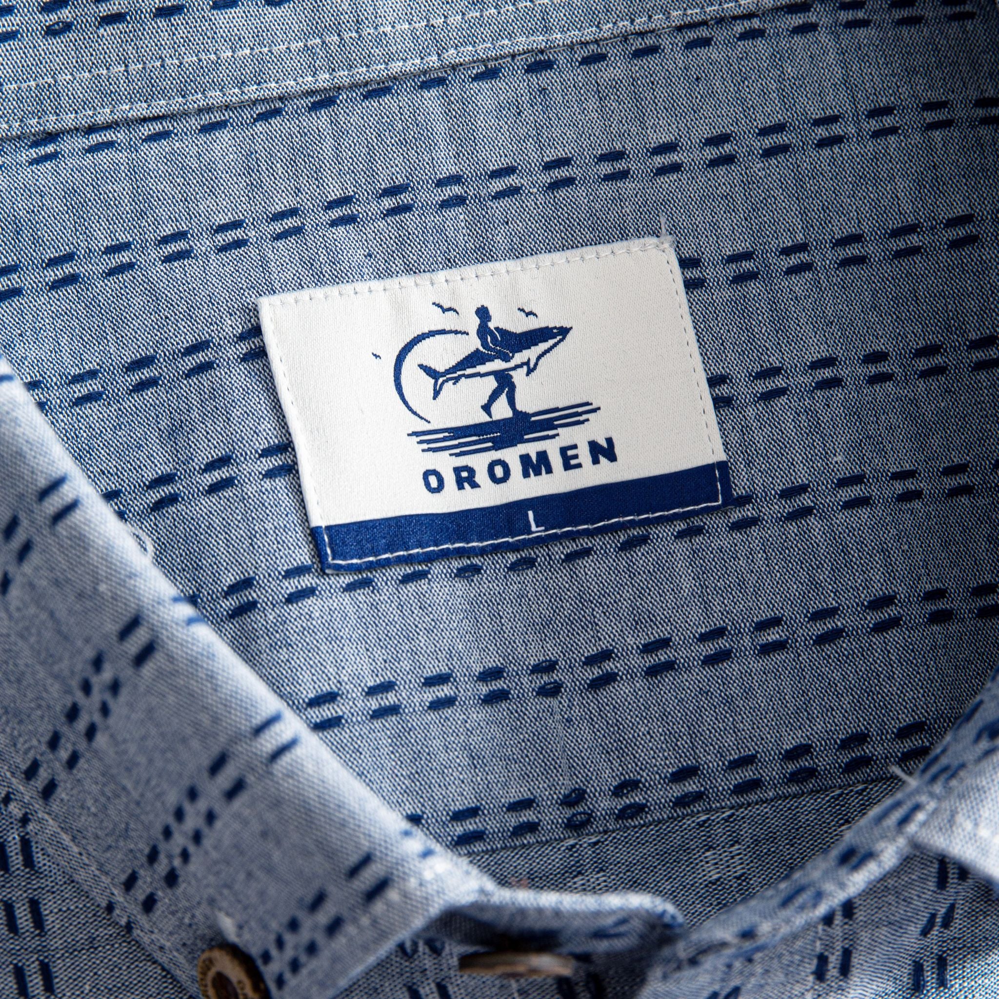 OROMEN Sashiko Chambray shirt branded woven label close-up inside collar, navy Sashiko stitch background