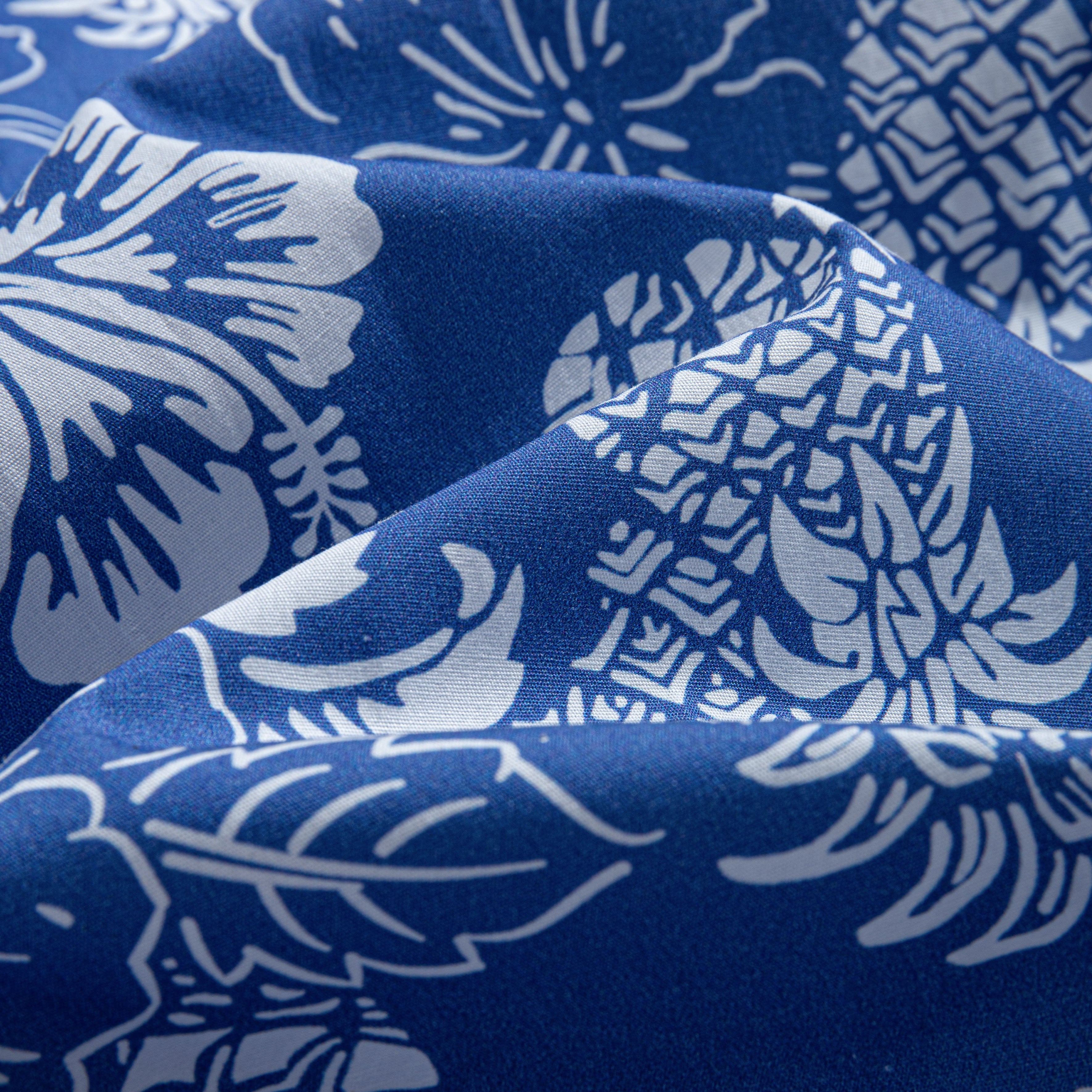 OROMEN Tropical Paradise shirt – draped fabric close-up showing navy blue Cotton-Tencel blend with white hand-painted hibiscus, pineapple and monstera leaf motifs, highlighting soft fabric drape and sheen