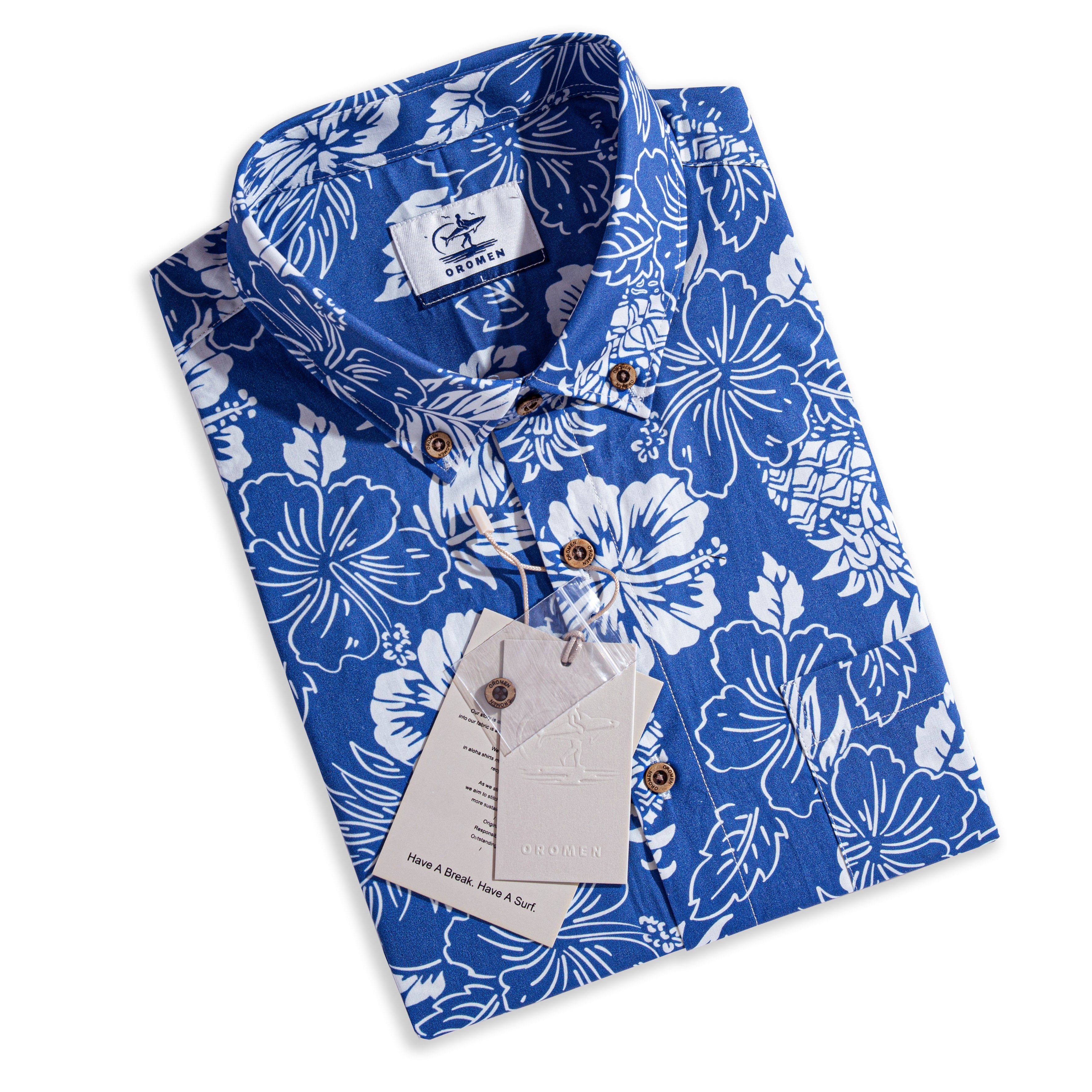 OROMEN Tropical Paradise Hawaiian Shirt – folded flat lay with OROMEN brand label and hang tag reading "Have A Break. Have A Surf.", coconut buttons, navy blue with white hibiscus and tropical floral print